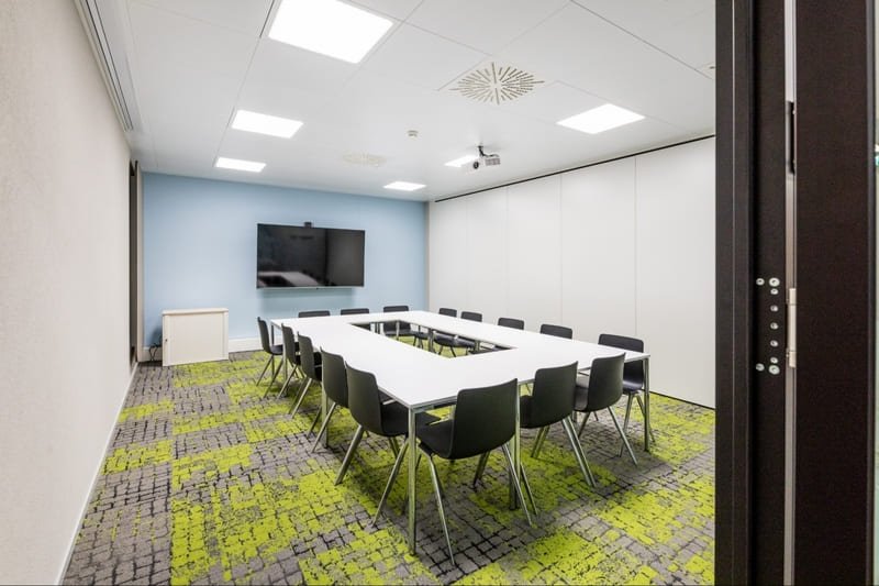 Konferenzraum mit rechteckigem Konferenztisch auf grün-grauem Teppichboden, ein Bildschirm befindet sich vor einer blauen Wand