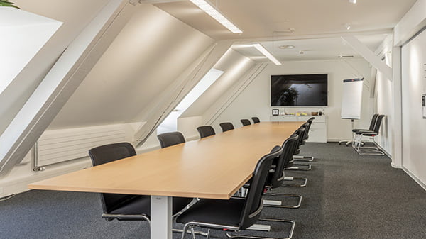 Meetingraum langem Tisch und jeweils sechs Stühlen links und rechts neben dem Tisch, an der Wand ist ein Bildschirm befestigt, rechts daneben ein Whiteboard
